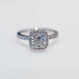 platinum ring with a 1ct cushion cut diamond as the centre stone and a halo of diamonds surrounding the centre stone as well as along the band.