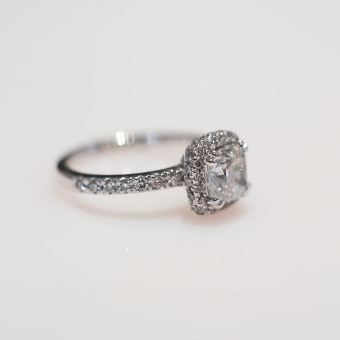 platinum ring with a 1ct cushion cut diamond as the centre stone and a halo of diamonds surrounding the centre stone as well as along the band.